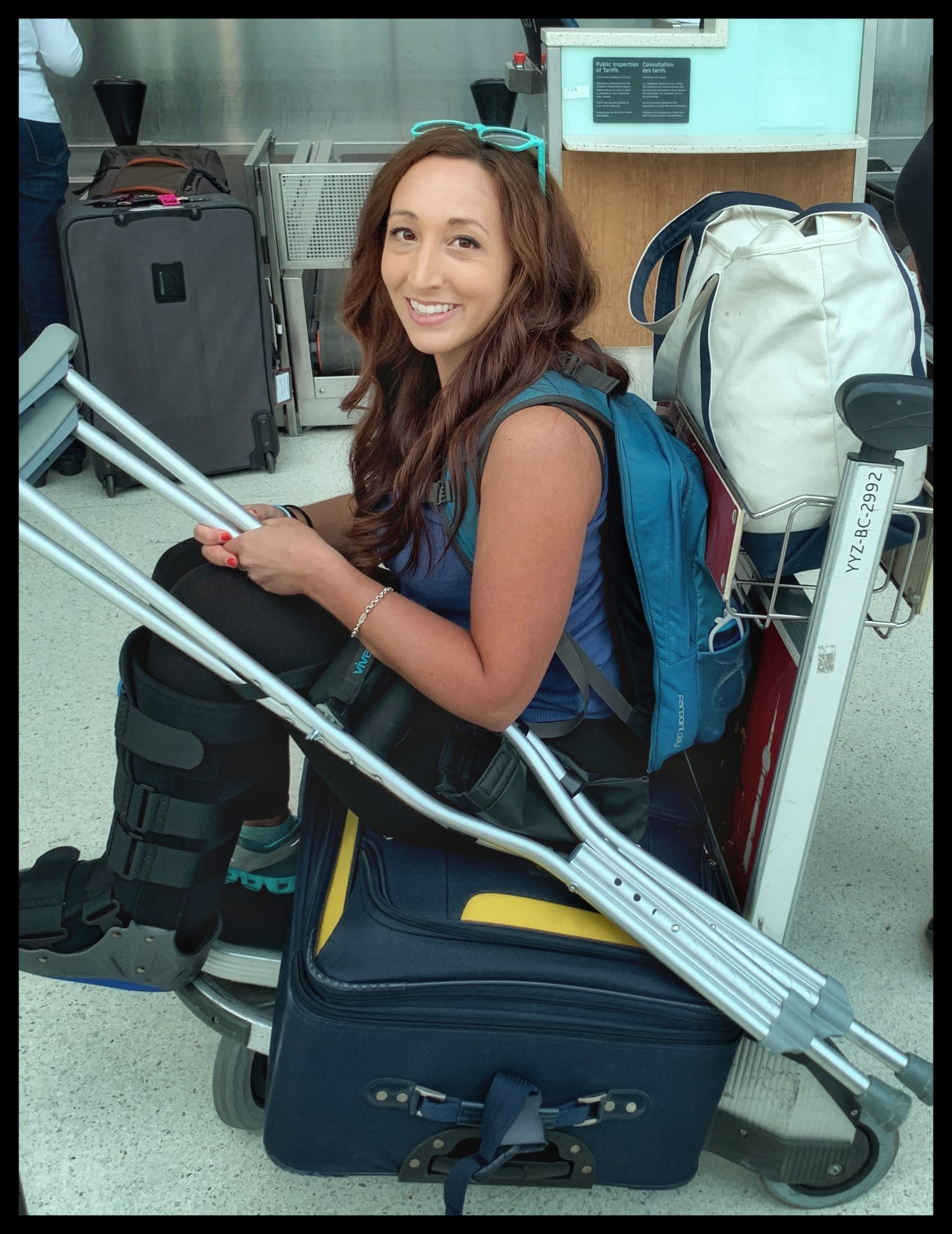 Girl sitting on a suitcase on top of a luggage cart with crutches