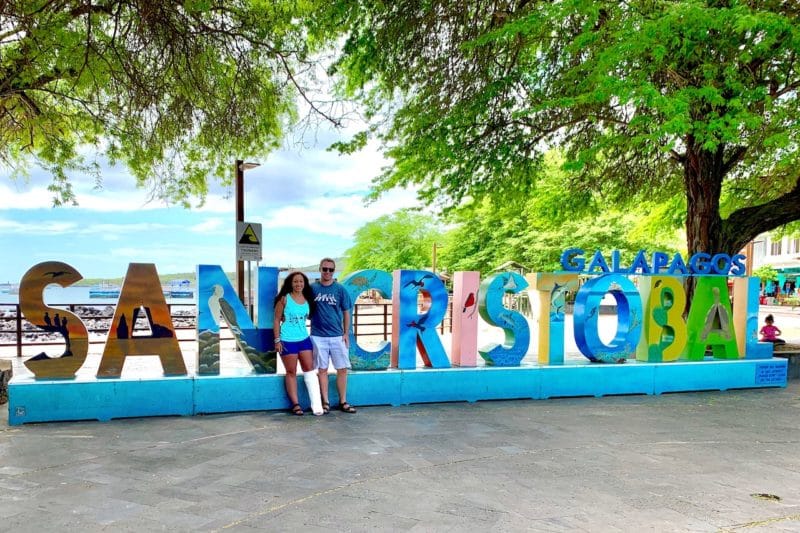 Couple standing in front of San Cristobal Galapagos Island sign. Girl with a broken leg.